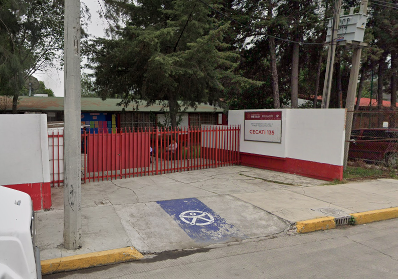 Fotografía en la que se muestra la entrada del plantel, conformada por una reja de color rojo por la que se puede ingresar a las instalaciones. Al lado derecho de la reja hay un muro blanco que tiene una placa que dice CECATI 135 y frente a la reja hay una rampa de acceso.